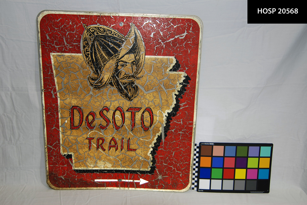 Metal painted sign with figure of Hernando de Soto, the outline of the state of Arkansas and words "DeSoto Trail," and double headed arrow to indicate direction. Red, gold, and black paint on sign is crackled and peeling off the metal backing. Found in collection during move of artifacts from Fordyce.