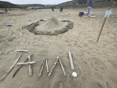 A sand sculpture of a small mountain surrounded by a moat with sticks in the foreground spelling out "TAM!."