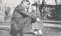 A black and white field of a man kneeling with a toddler in the grass. He is wearing a suit and facing her with arms outstretched. The little girl has shoulder length hair and is wearing a long jacket and a hat. They are facing each other grinning. There is a building in the background. 