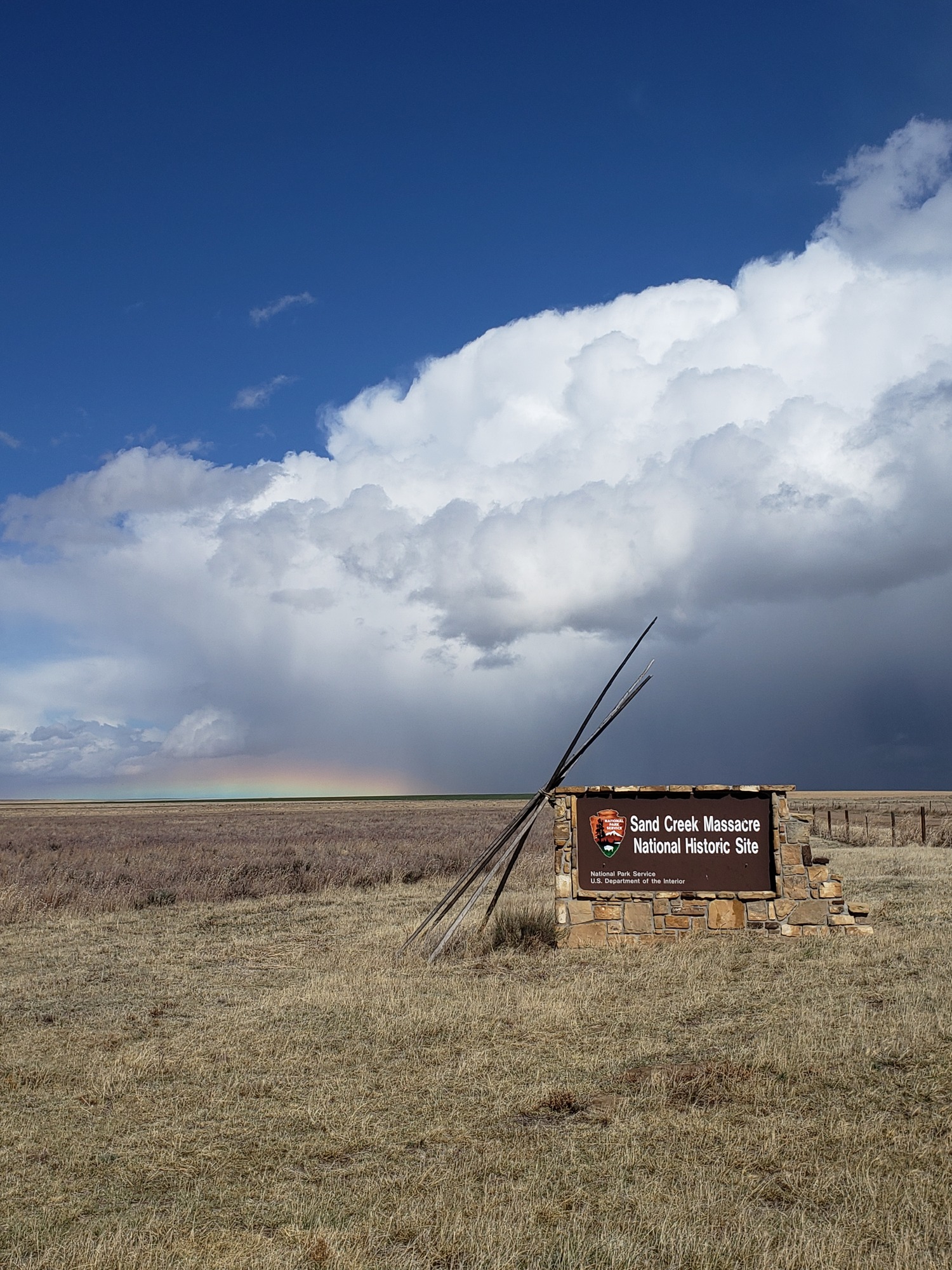 A picture of the Sand Creek Massacre entrance sign. Behind the sign a short rainbow sits on the horizon with storm clouds in the distance. 