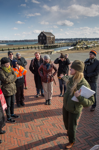 Group of people stand in a semicircle facing a female park ranger who is holding papers in one hand and has other arm extended as she speaks to the group. They are outside standing on brick pavement with waterfront visible behind them. 
