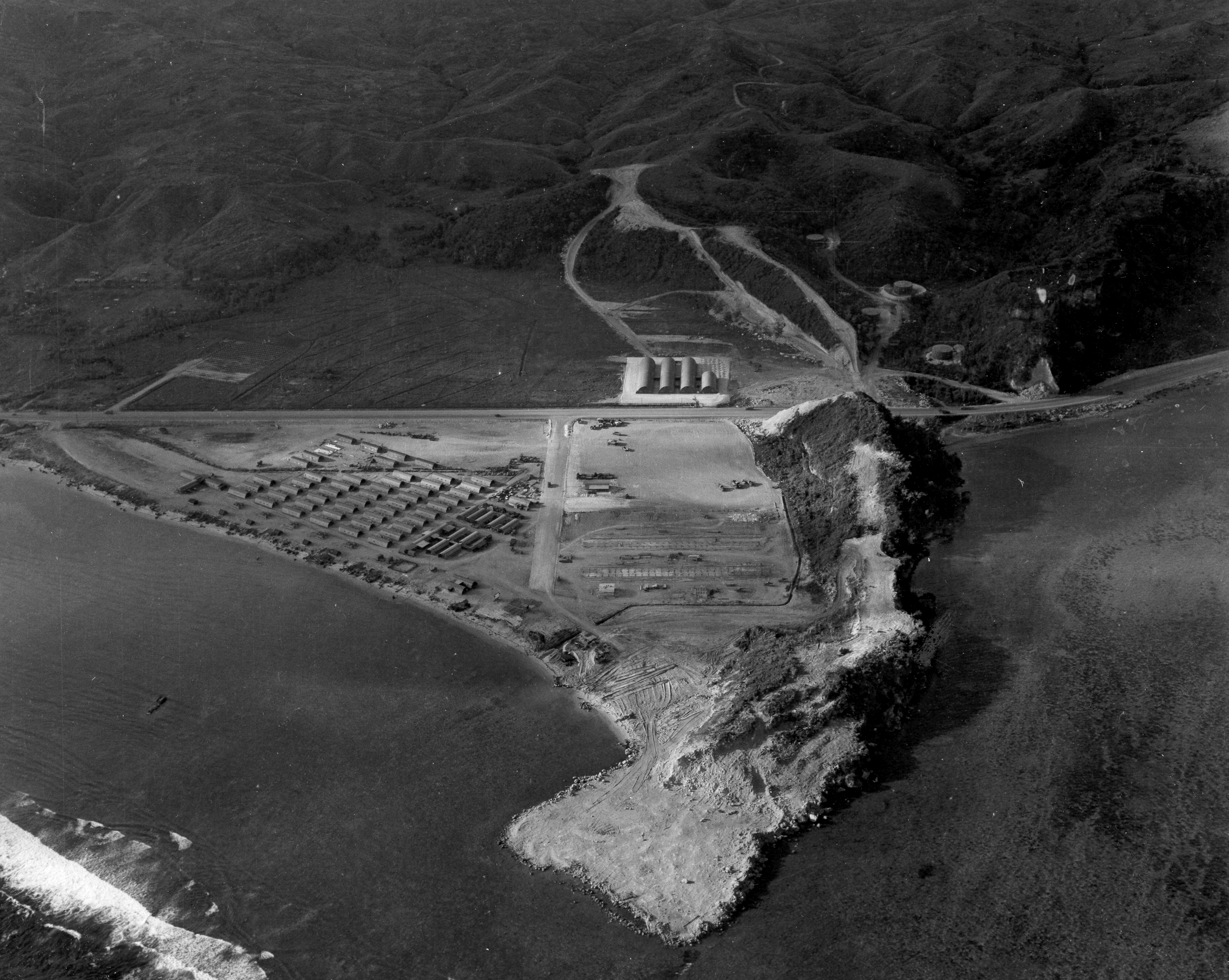 Aerial view of a point of land sticking out into the ocean. The ridge on the right has been heavily deforested. In the plain to the left, an orderly military base has been laid out. A road runs through the plain behind the point.