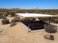 Two picnic tables under a small shade structure. A fire pit is next to the shade structure and desert vegetation grows around. 