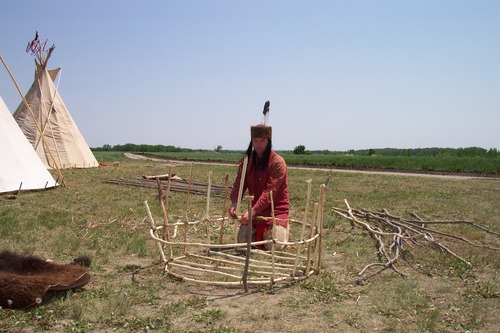 A man in native dress tying sticks into the shape of a boat in a grassy field with canvas tipis beside hime