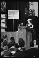 A seated audience watches a woman who speaks from behind a tall podium with two microphones, next to a sign that says 