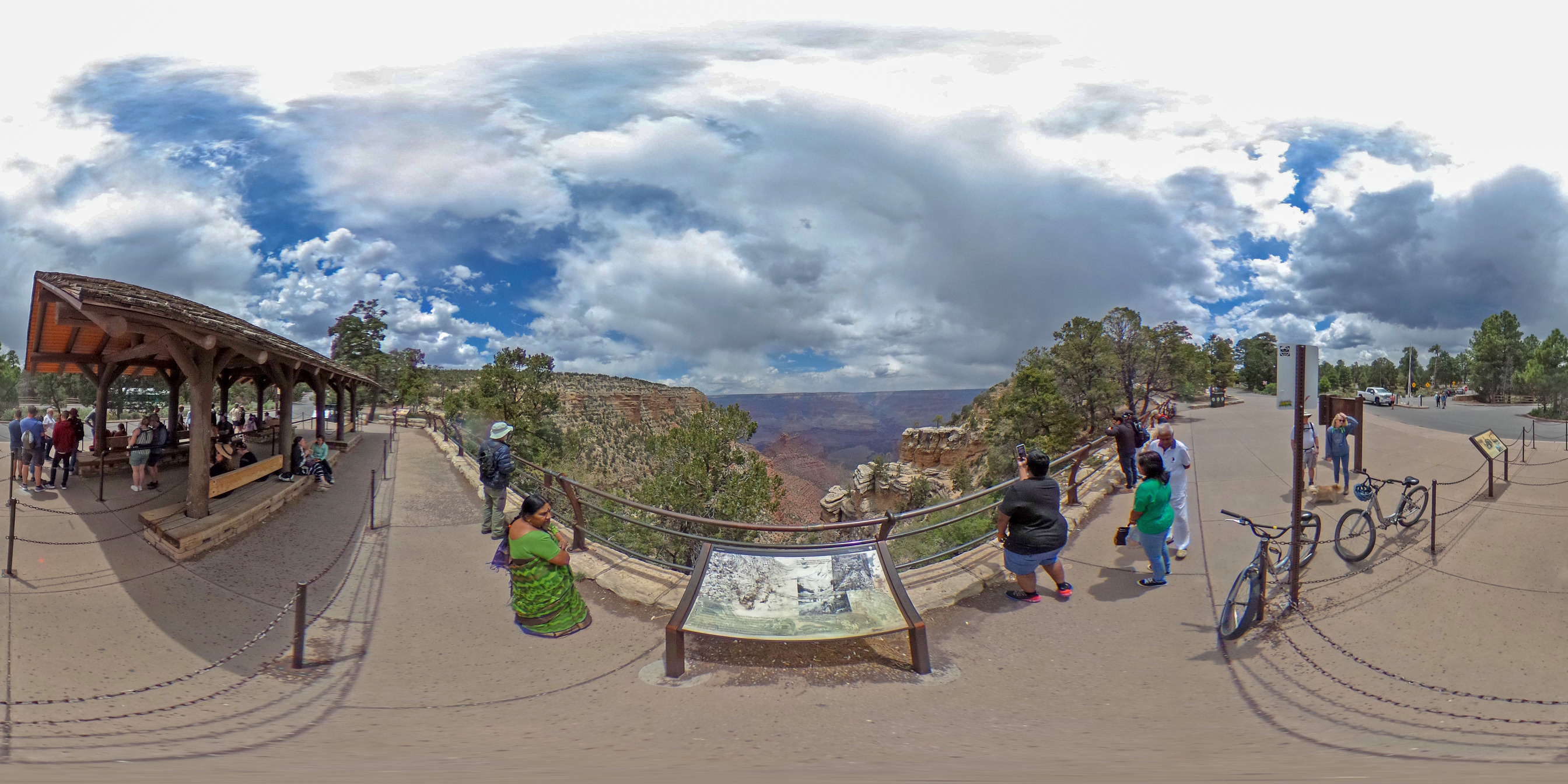 A 360 spherical panorama showing a faded wayside panel in the center with several people on either side looking into a canyon landscape. On the left is a bus stop shelter. 