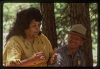A woman in a Native American attire stands next to a seated male park ranger in uniform. 