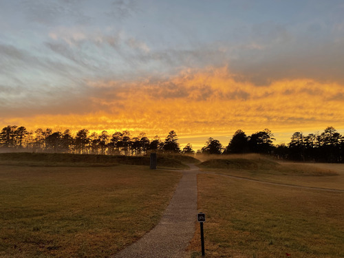 A bright orange sunset silhouettes a line of trees behind an earthen fortification covered by a thin line fog.