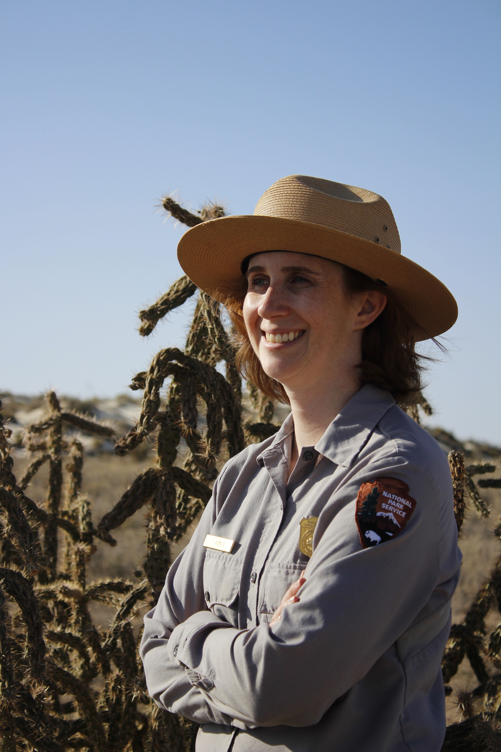 A woman wearing the national park service uniform poses for a photo on the border of the White Sands dune field. Cacti and desert shrubbery extend into the distance meeting the base of white sands dunes. The dunes are freckled with shrubbery and meet a clear light blue sky on the horizon.