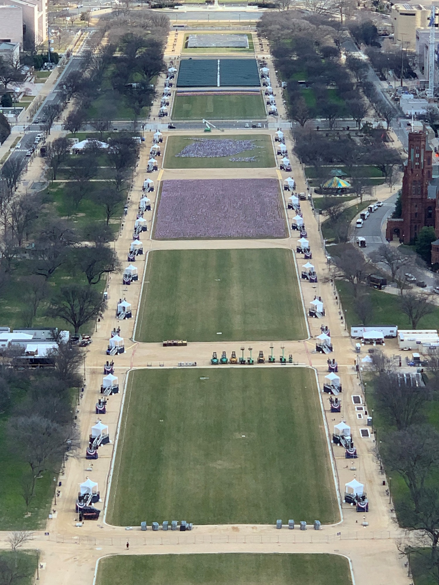 An aerial view of a sections of lawns filling in with state flags. The lawns are lined with wide pathways covered with spaced tents. 