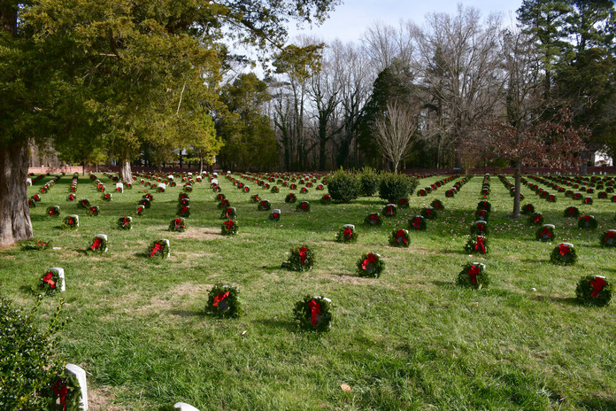 Hundreds of green wreaths with red bows lean on white marble headstones surrounded by green grass. 