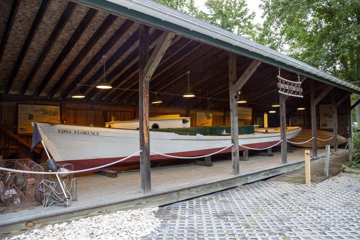Covered pavilion displaying historic white boats, including one named “Edna Florence", and informational panels along the back wall.