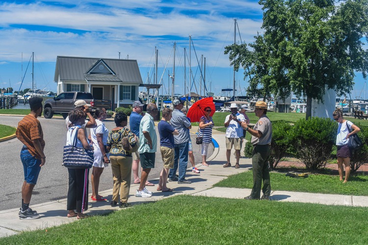 An NPS Ranger can be seen giving an interpretive talk to a group of 13 guests standing on a sidewalk at historic Long Wharf in Cambridge, MD. Three bushes, a tree, a home, and many docked boats can be seen in the background.