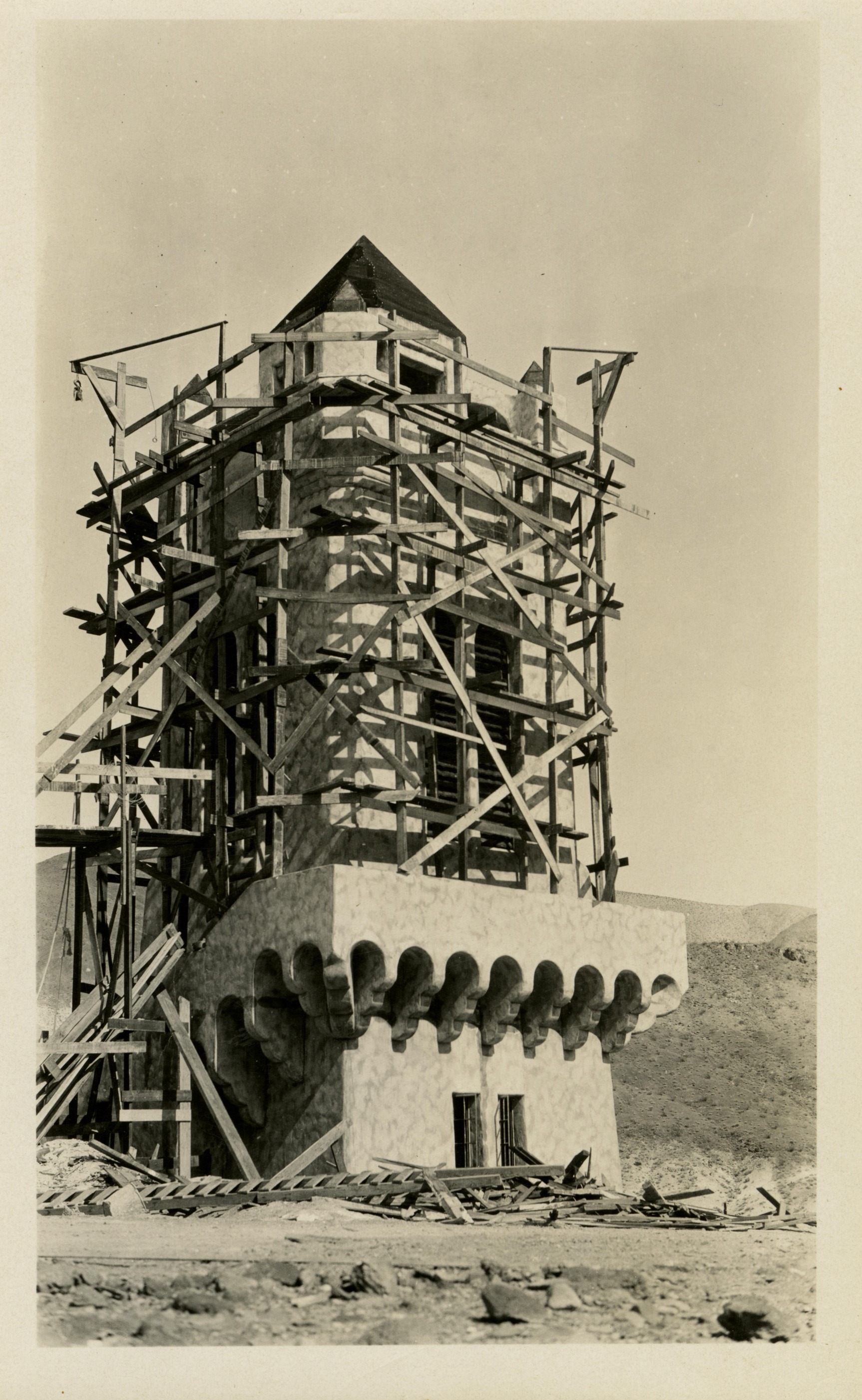 This is an historic black and white photograph from the Scotty's Castle Historic Photograph Collection, Death Valley National Park close-up of square tower encased in scaffolding. Base with wrapped stairway across front only section not scaffolded. Lumber construction debris around base. Steeple roof line peaks above scaffold.