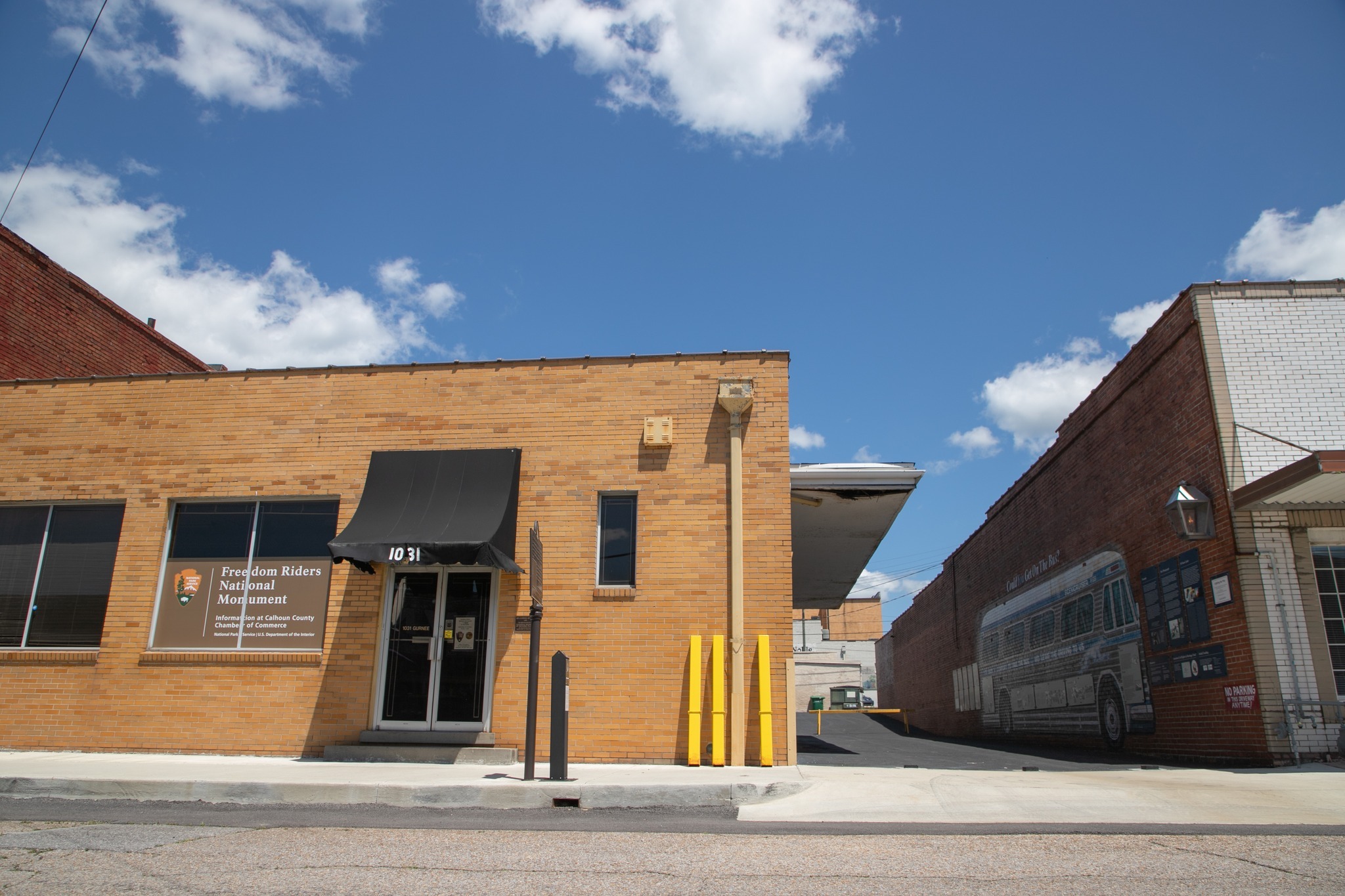 Large NPS sign in window of rectangular brick building that faces a city street and has an alley alongside. Building across the alley bears a mural of a bus