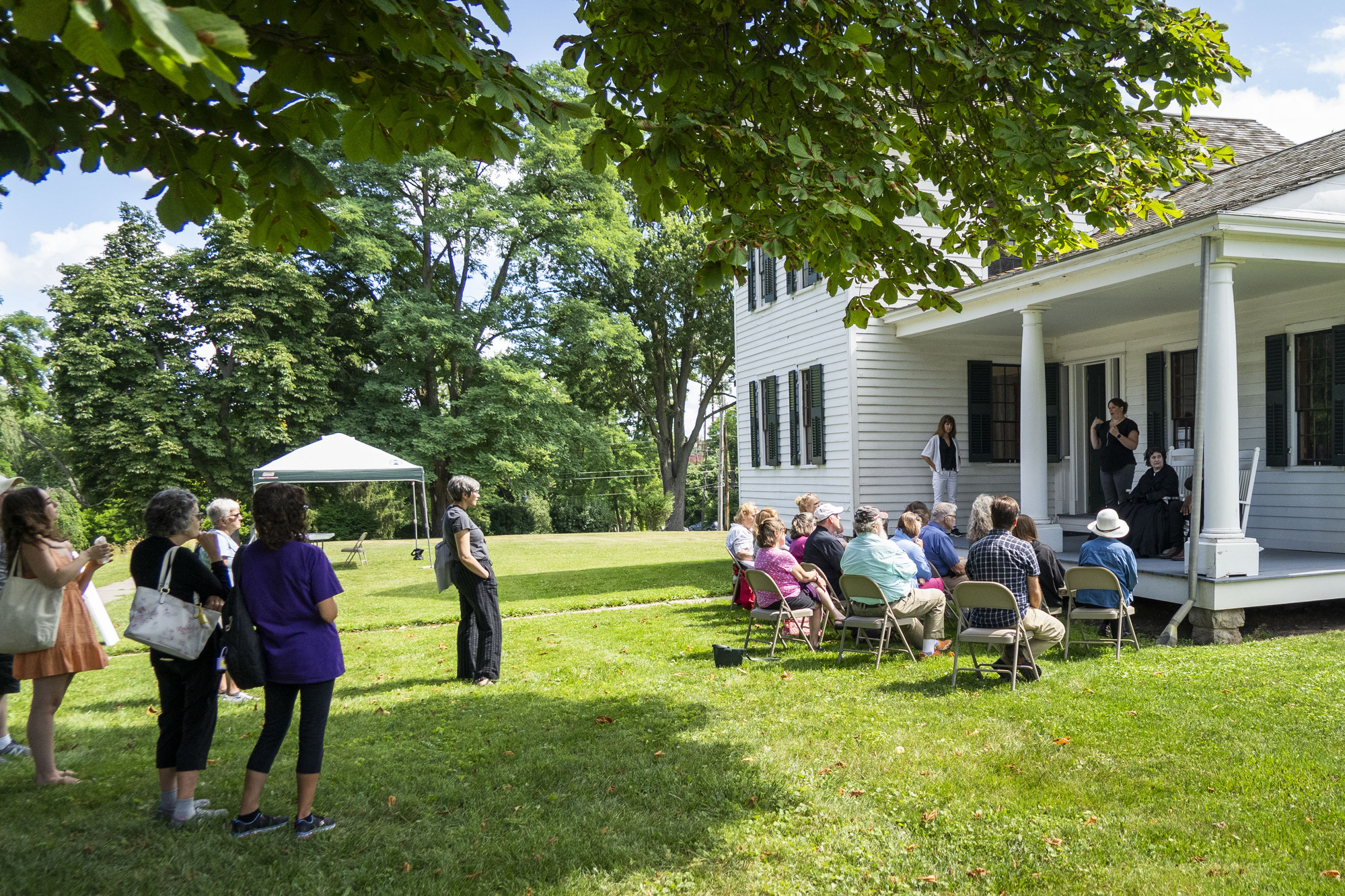 A woman and man wearing historical clothing sit in rocking chairs on the porch of a house. A group of people is gathered in chairs in front of them.