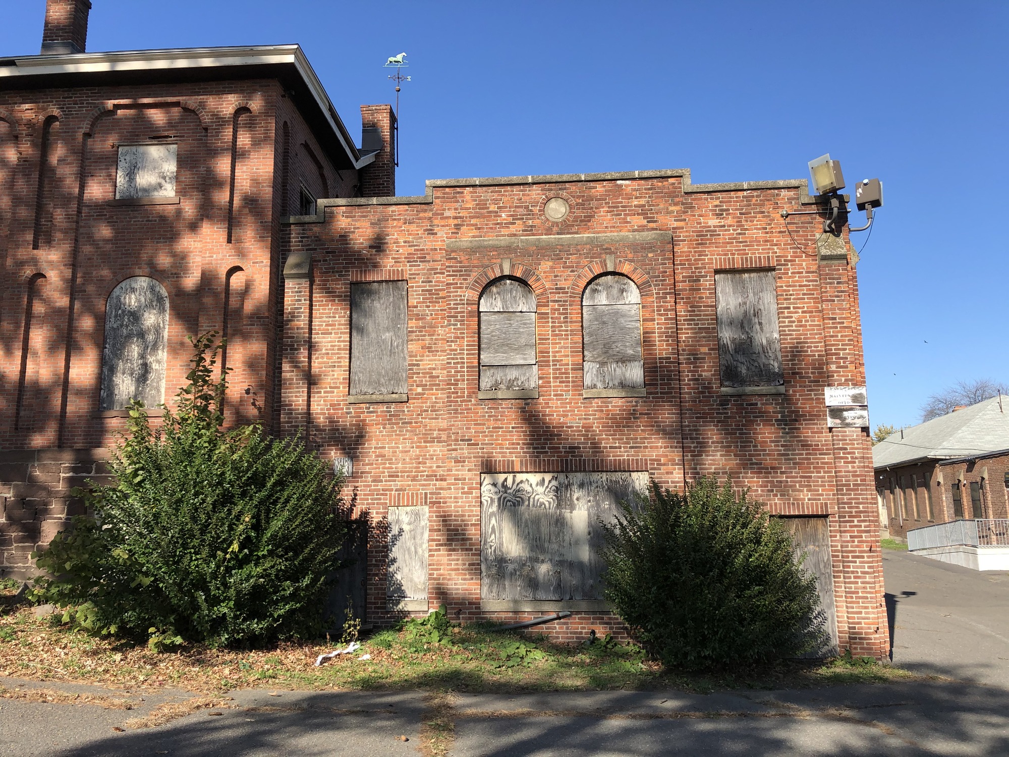 Brick building with several boarded up windows and two overgrown shrubs.