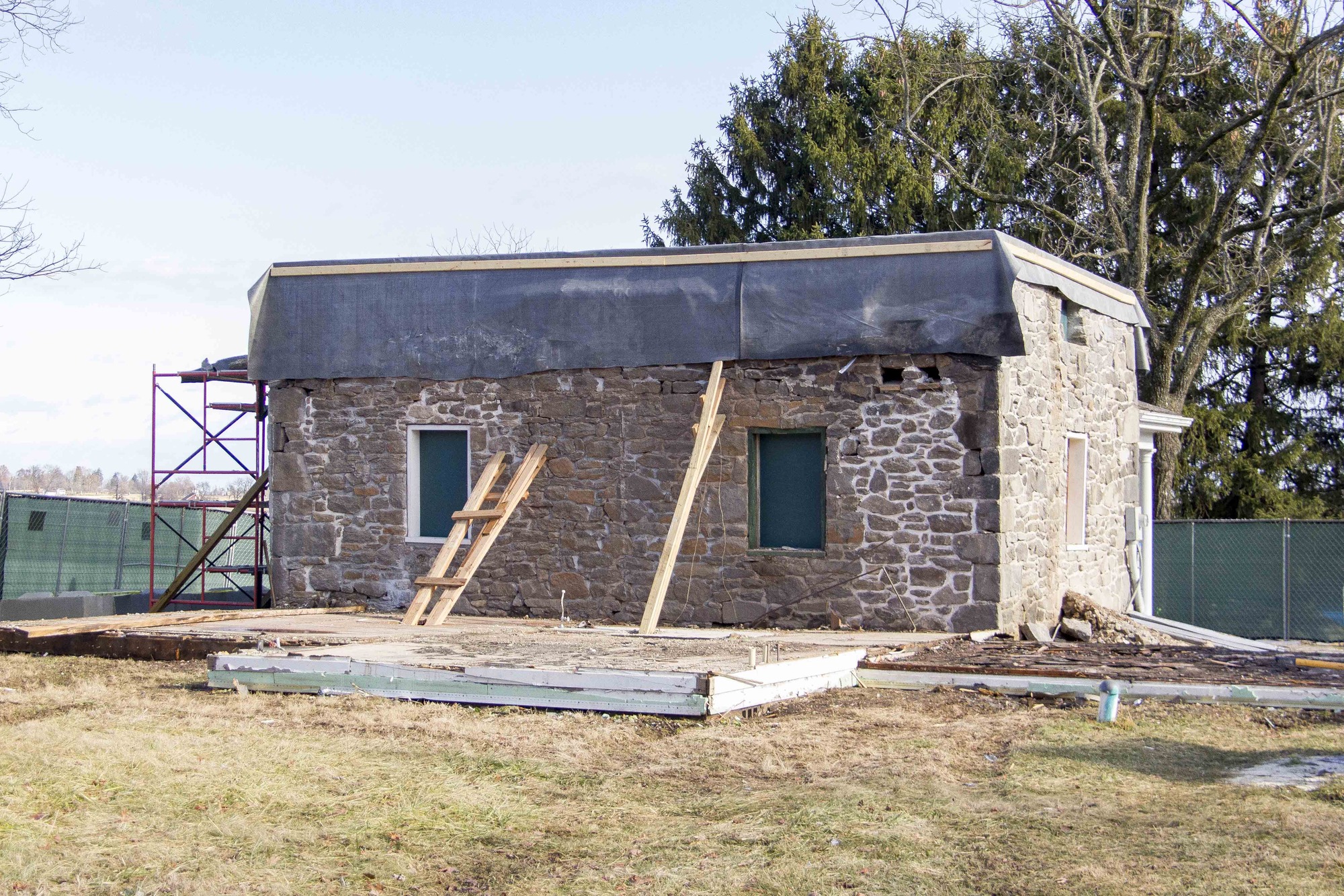 The rear view of the Warfield house after the selective demolition process. The two historic windows have been repaired. There are two wooden stabilizers leaning against the masonry on the back of the house. There is a black tarp protecting the roof of the original house structure. The red construction element is on the left side of the house. 