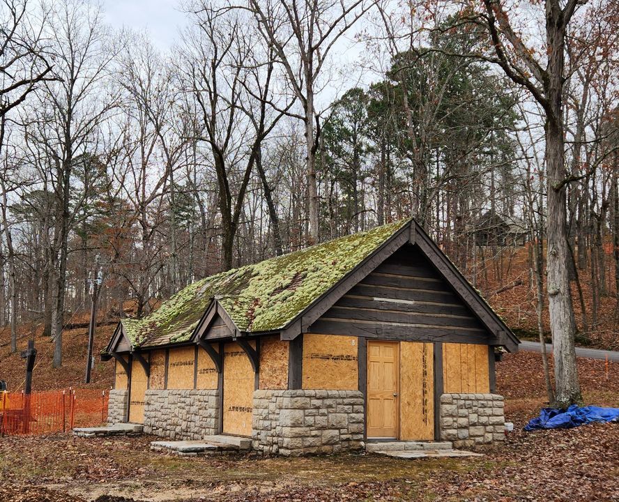 A stone and wood building with a moss and lichen covered peaked wood shingle roof with peaked entry covers stands in a forest with no leaves and cloudy sky visible through the trees. The lower half of the building is made of coursed cut stone except where two doorways in the front and one on the near end interrupt the stonework. The roof is held up by regularly spaced wooden beams in between doorways and windows, and the end wall above the doorway is made of horizontal wooden beams with mortat in between. The doors and windows have been removed and replaced with plywood.