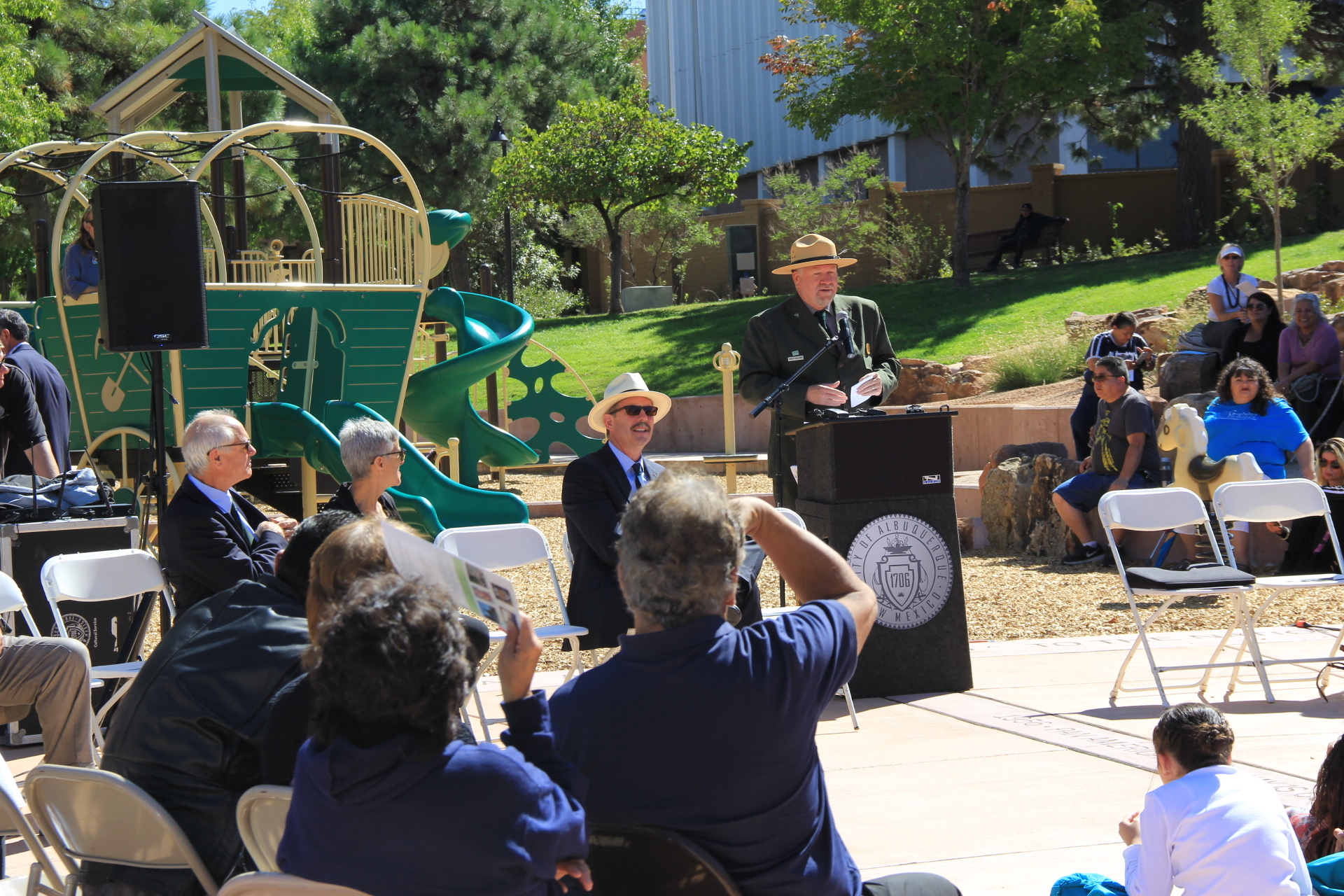 A man in a ranger uniform speaking at a podium during an outdoor event with people sitting and listening, and a playground in the background.