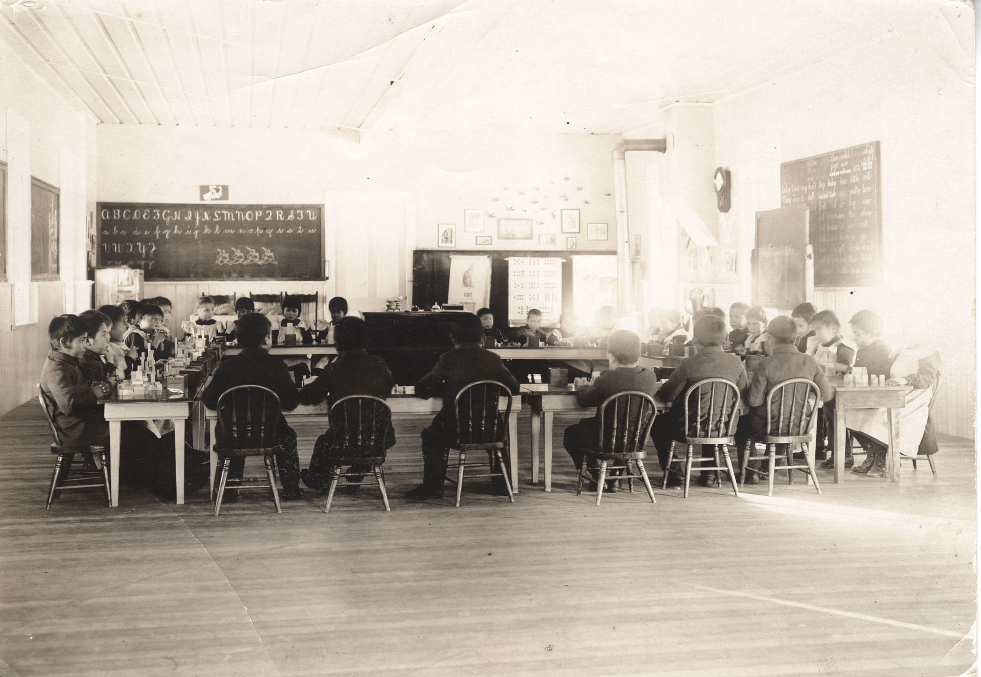 Black and white photograph of children sitting at tables in a plain room with posters and chalkboards on the walls