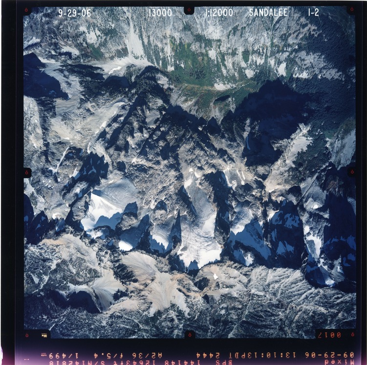 Color aerial photograph of glacier and snowfields on mountain peak with forested valleys below.