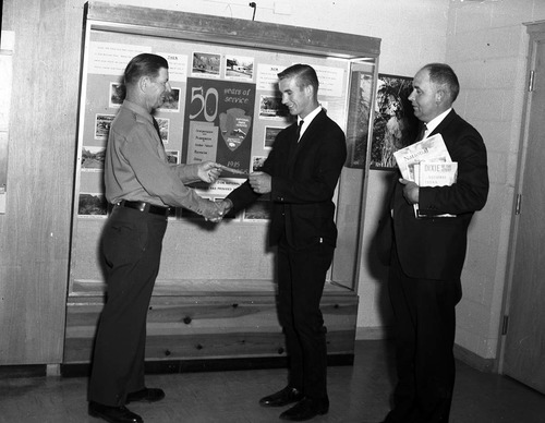 Superintendent Warren F. Hamilton presents Creed Fraehner with award in front of Fraehner's winning poster for the National Park Service 50th Anniversary, inside the Mission 66 Visitor Center and Museum. Jim Clarke stands by with gift of books for Fraehner.