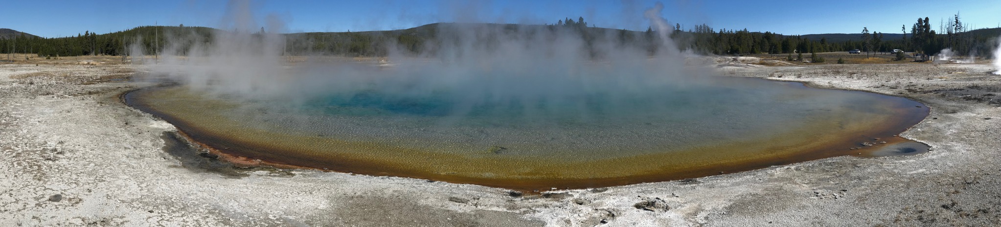 Steam rises from a round, blue hot spring with a rim of yellow and orange colors sitting amongst a white field
