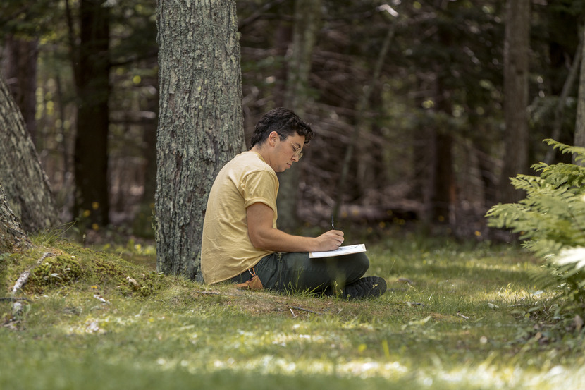Artist-in-Resident seated on grass, sketching in a notebook in a forest.