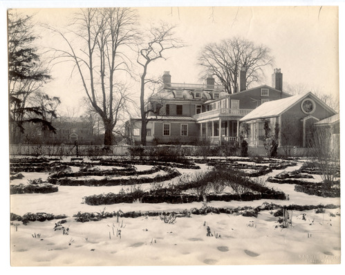 Black and white photograph of formal garden in the snow with large Georgian mansion in the background.