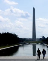 Two people stand in front of a reflecting pool leading to the Washington Monument. 