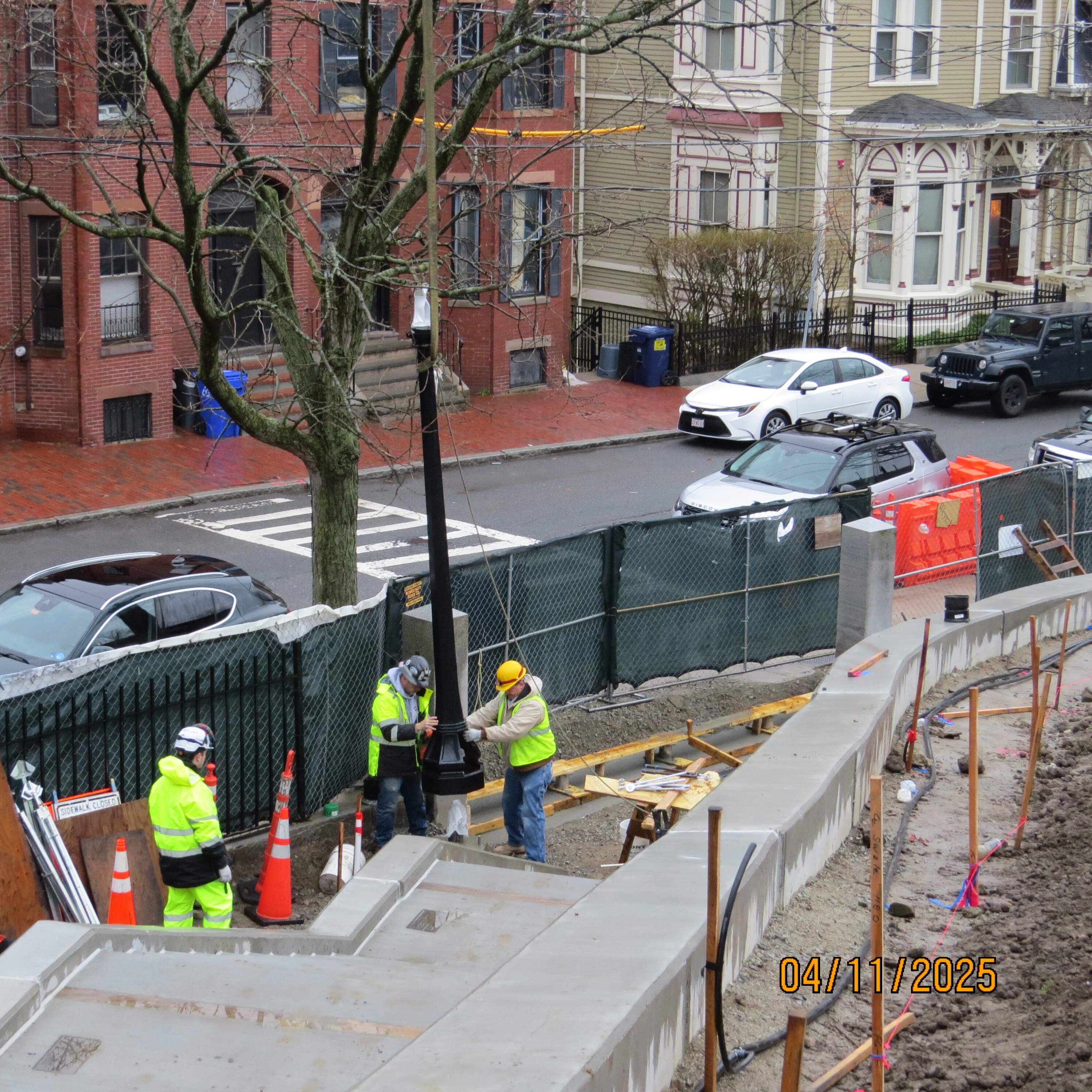 Workers lowering a black light pole into place along a concrete staircase that runs along the side of a hill. 
