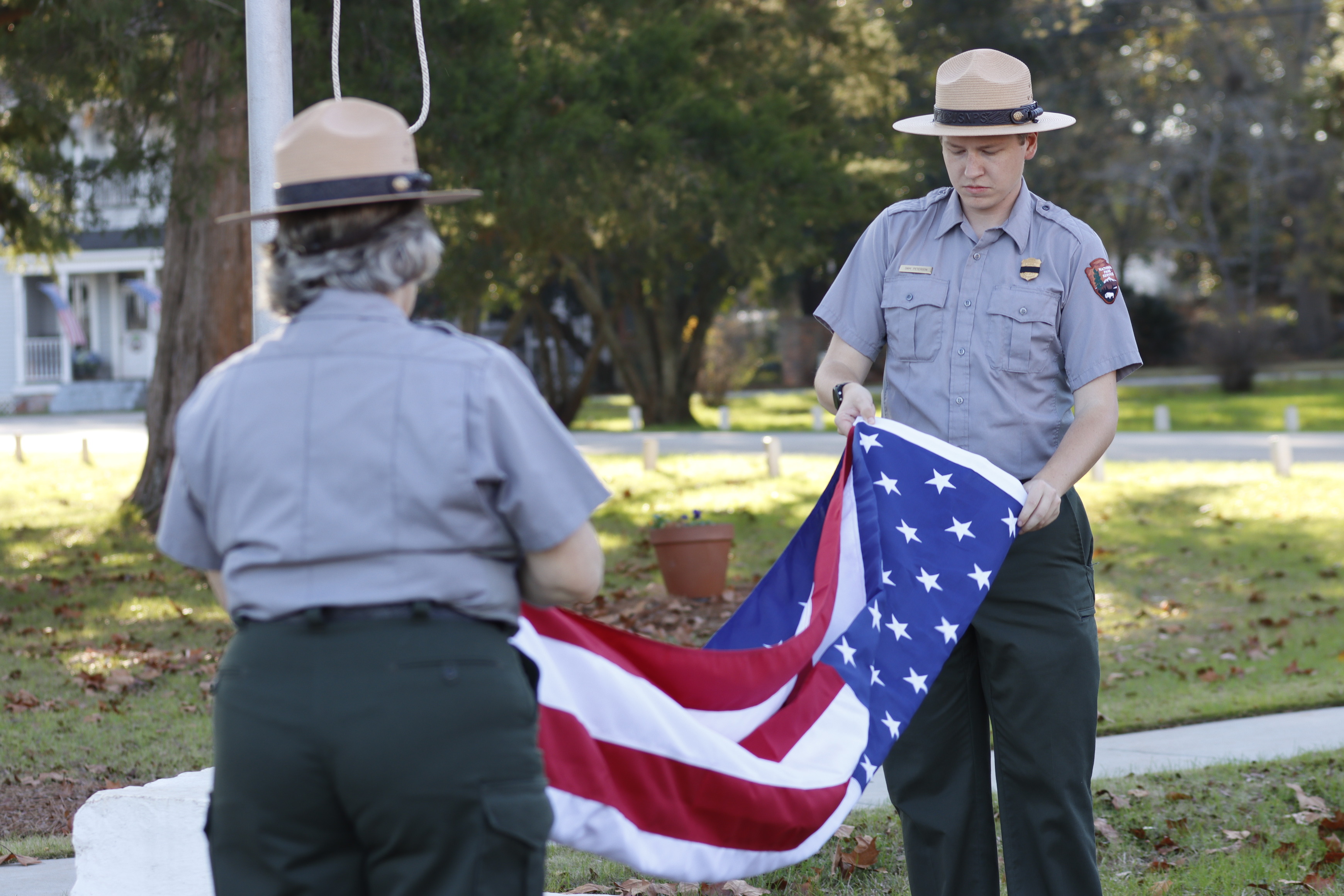 Park Rangers Sam and Mary Anne fold the American flag together. Both are dressed in their National Park Service uniforms, concentrating as they perform the traditional folding ceremony. The scene is set outdoors, with the flagpole and a backdrop of trees.