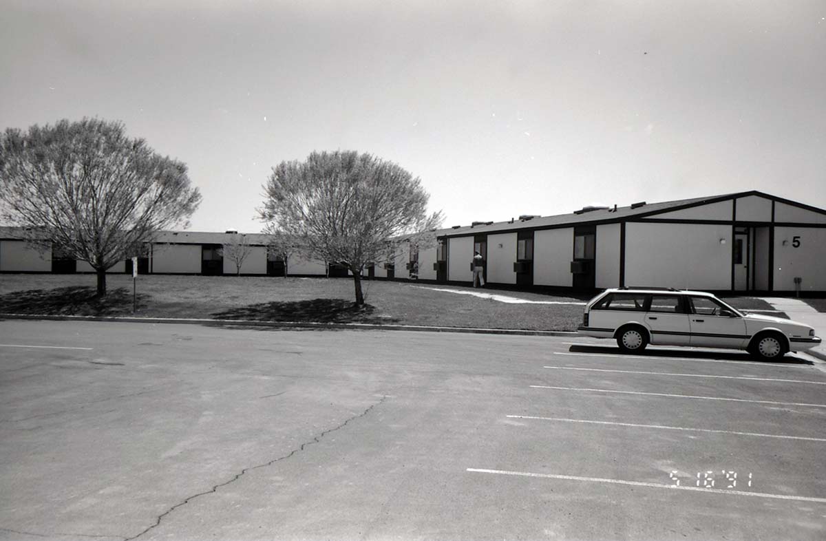 Barracks and parking area with man walking on sidewalk. Building number 5. [Image possibly for comparative housing study]