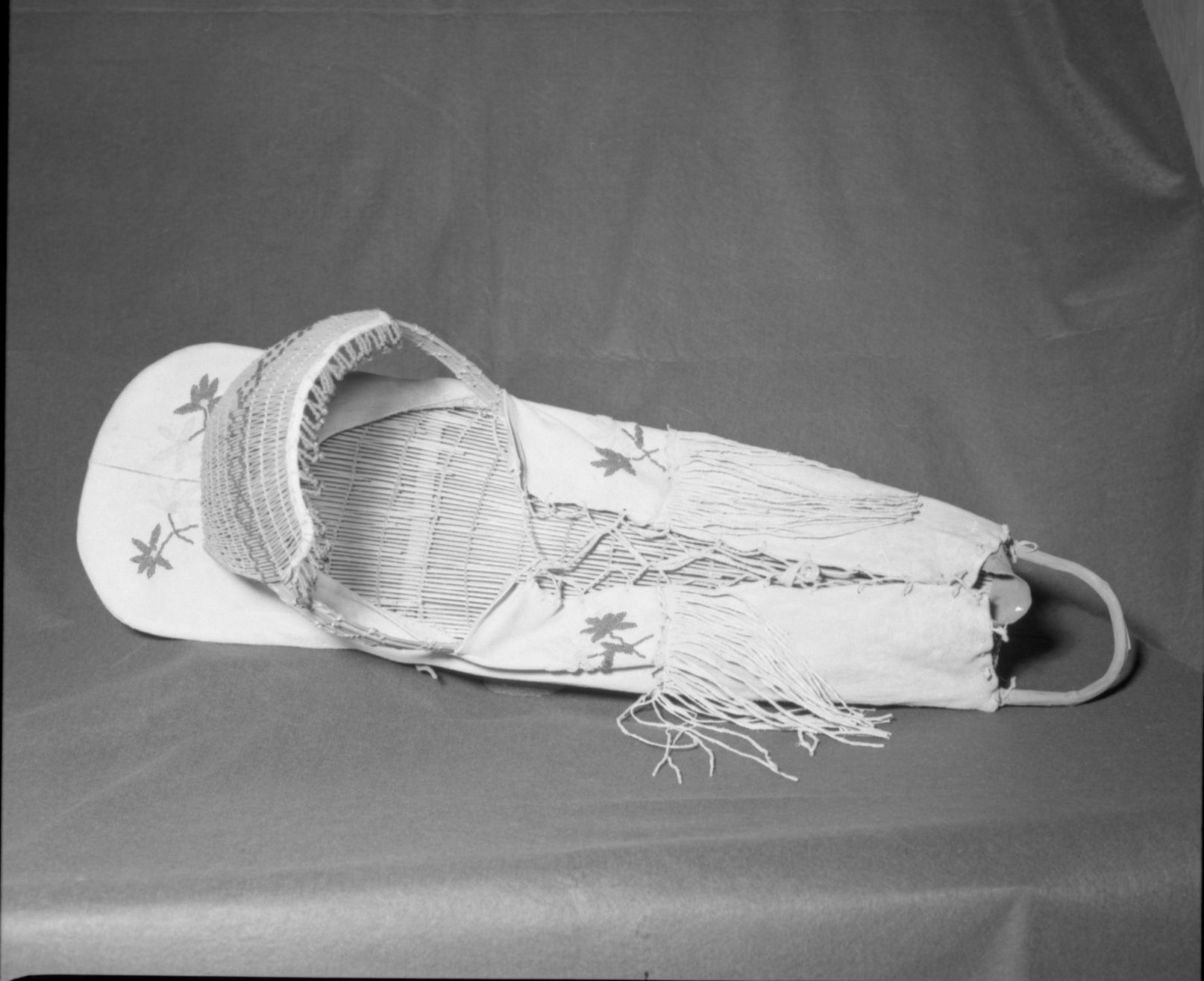 Mrs. Donald Segerstrom Collection. Object: Paiute cradle basket. Dimensions: 39" length. Materials: Willow, buckskin, glass beads, cloth, cord. Technology: Backboard plain twined, up to right slant of turns, hood diagonally twined with up to right slant of turns, flowers and leaves in modified raised lazy stitch, top seams of buckskin cover appear to be sewn on machine. Photographed by Michael Dixon. Catalogued by Craig Bates and Barbara Beroza. Yosemite Collections, National Park Service. 1/83.