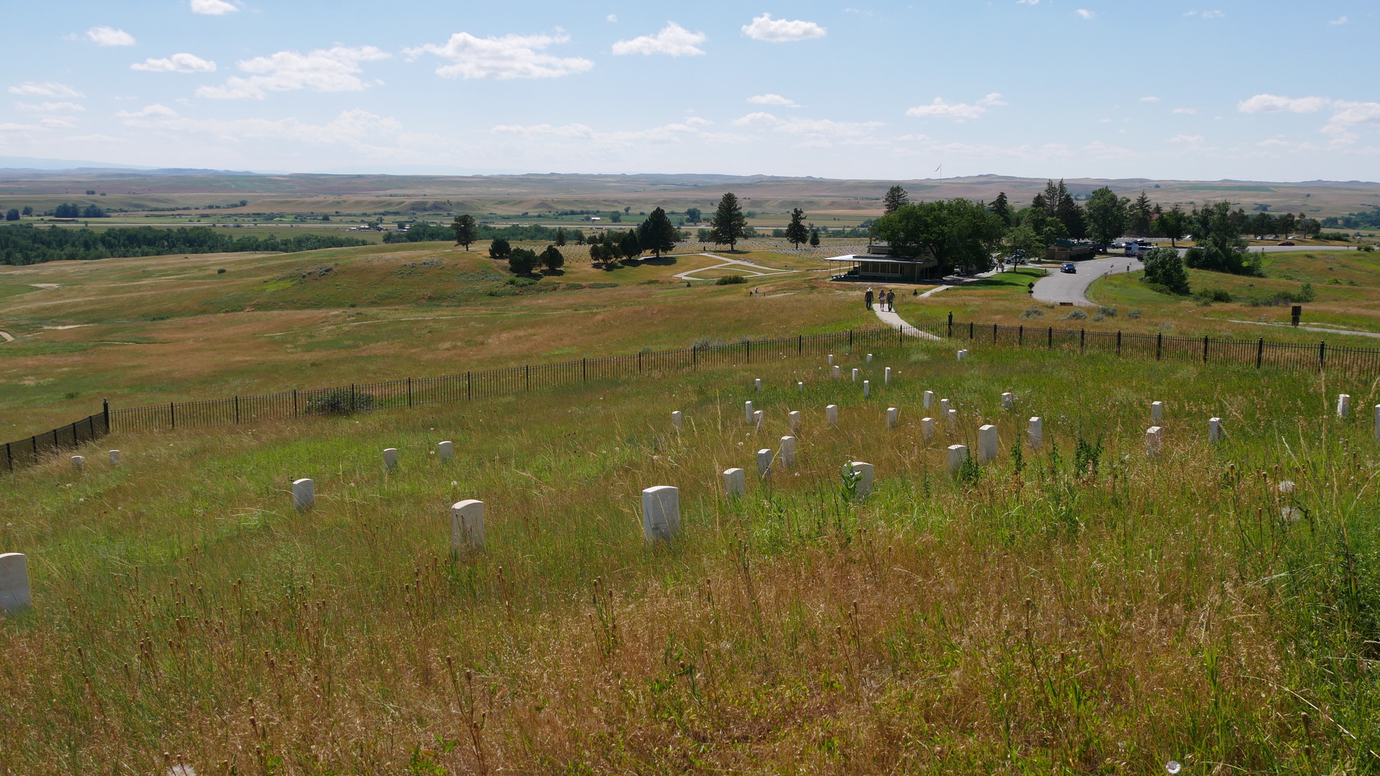 A metal fence encloses a sloping area that contains tall grass and scattered marble headstones. In the background and across gently rolling grassy hills, the visitor center building is to the right of the national cemetery, which contains uniform rows of grave markers and scattered trees.