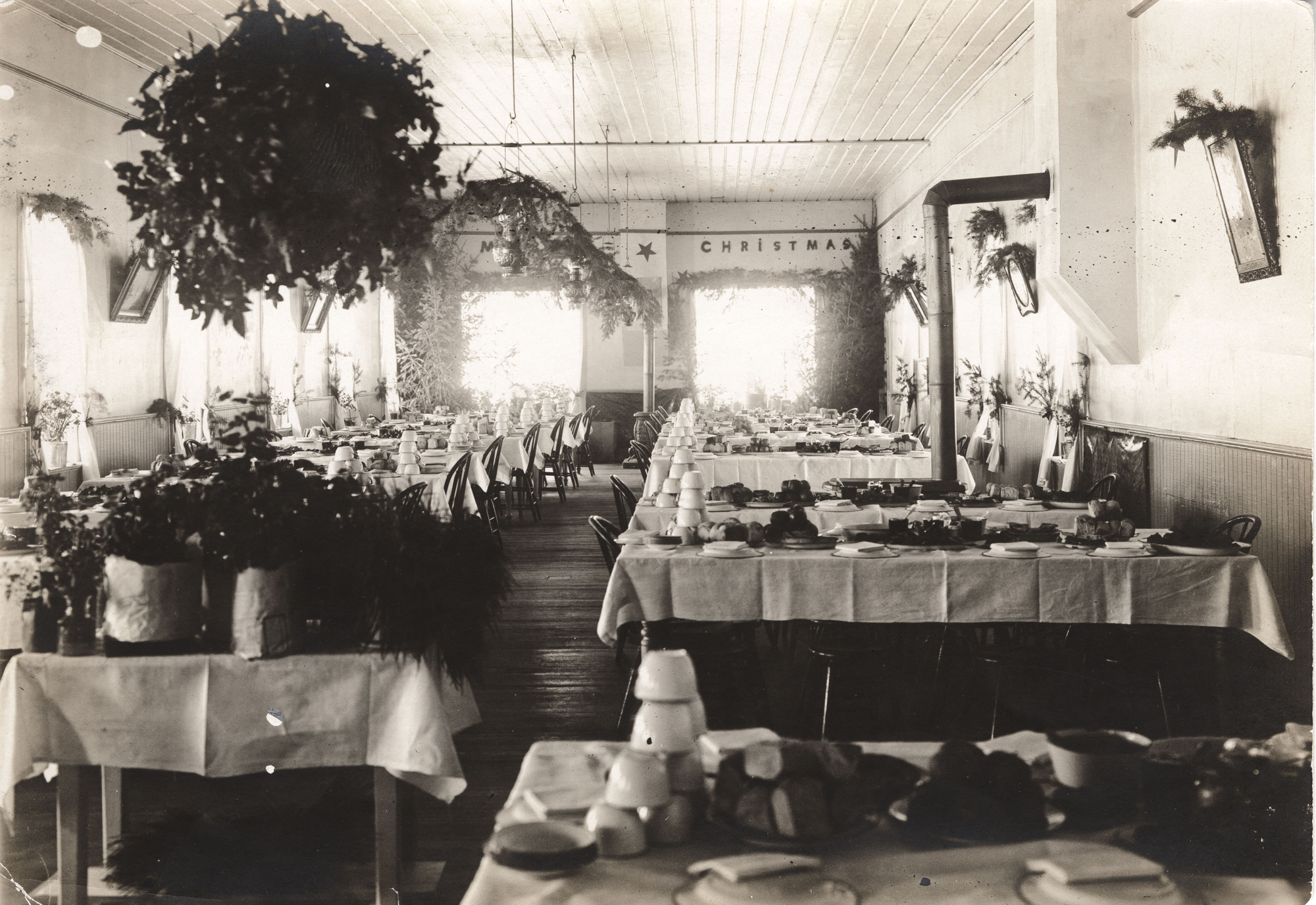Black and white photograph of a plain room with several rows of tables set with dishes and food with flowers and plants decorating the room