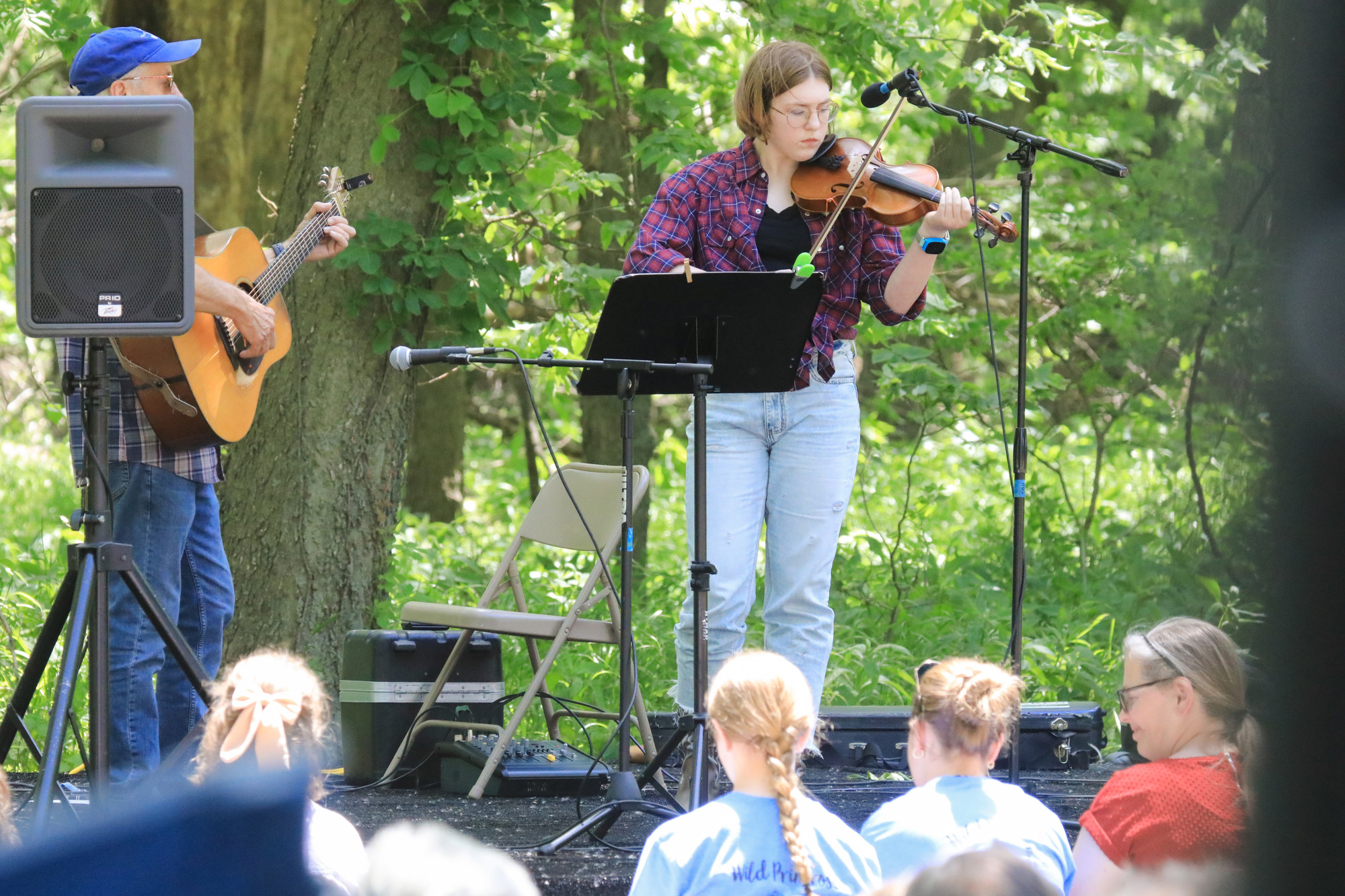 Young lady wearing blue and red plaid shirt and faded blue jeans plays violin in front of a seated crowd. She's accompanied by a man in a plaid shirt with a guitar.