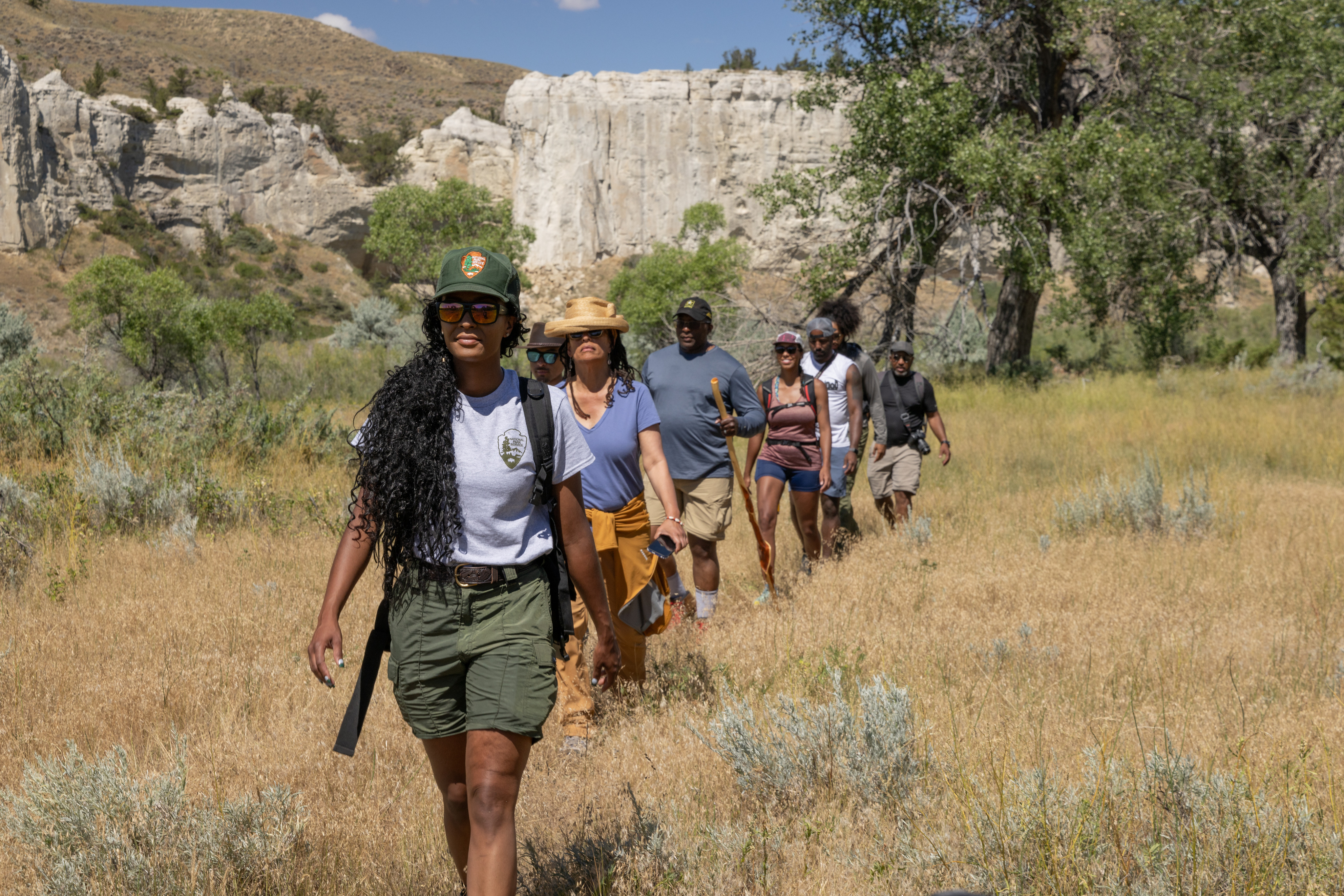 Eight people walking in single file on a hike through scrubland with white cliffs and trees in the background