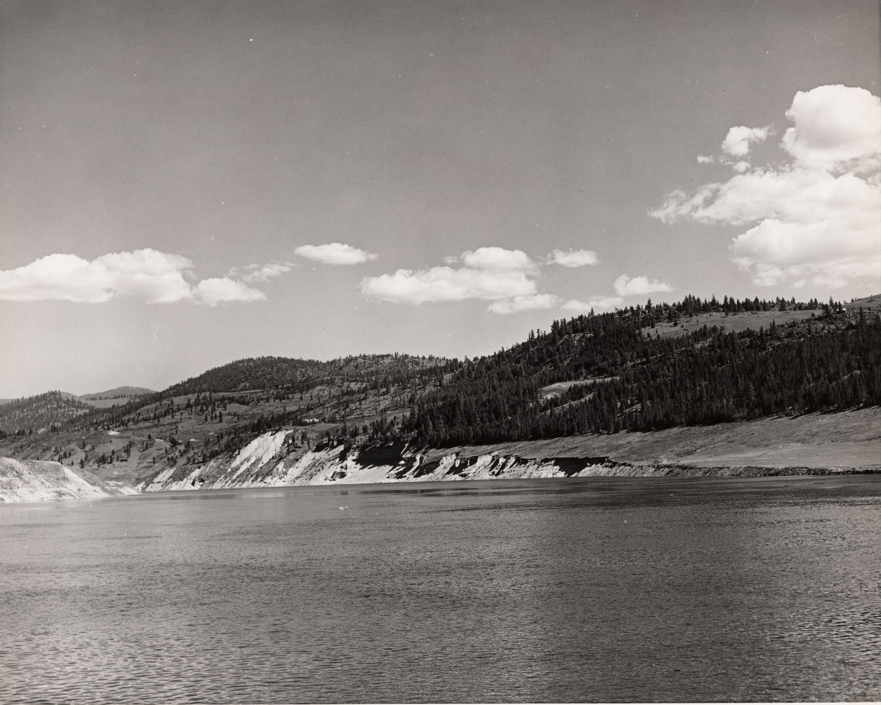 Black and white photograph of a large body of water with a hilly, partially forested shoreline