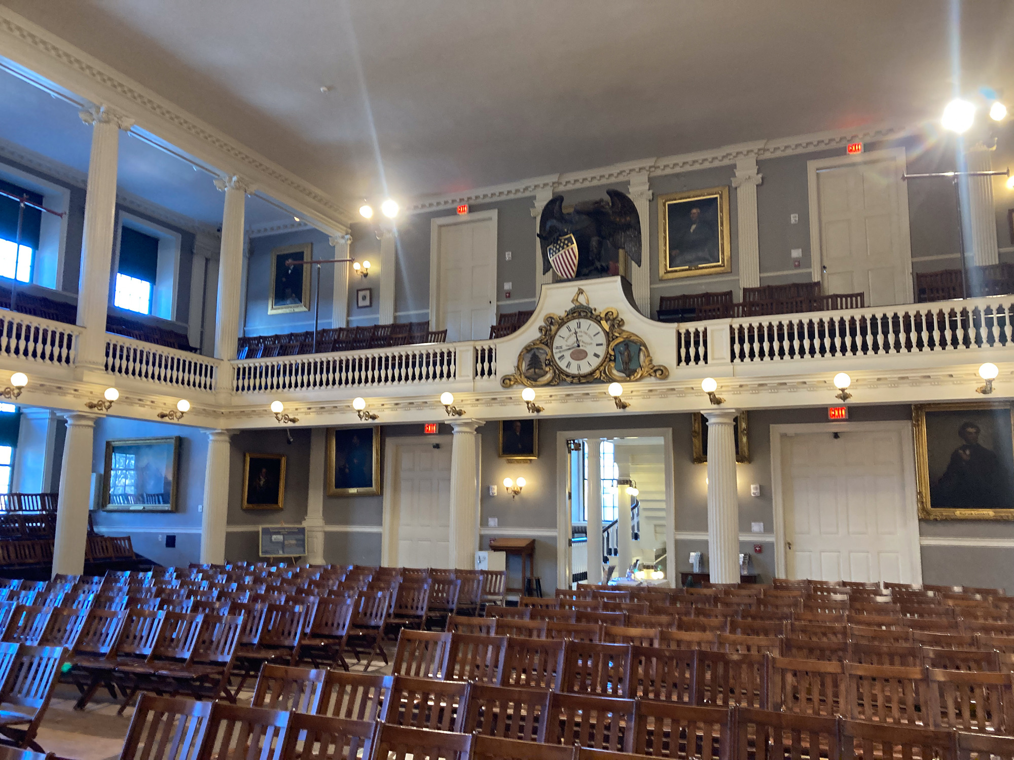 Interior view of back side of the Great Hall in Faneuil Hall, on the floor are brown wooden chairs that are connected. On the back wall is three big doors, the middle one is open. Above it is an ornate clock on the balcony railing. It has a big white clockface with gold gilding around the edges. Above it is a brown eagle holding a crest with red, white, and blue on it. 