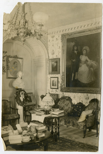 Black and white photograph of the corner of a 19th century parlor, featuring paintings and a bust, floor crowded with tables and chairs.