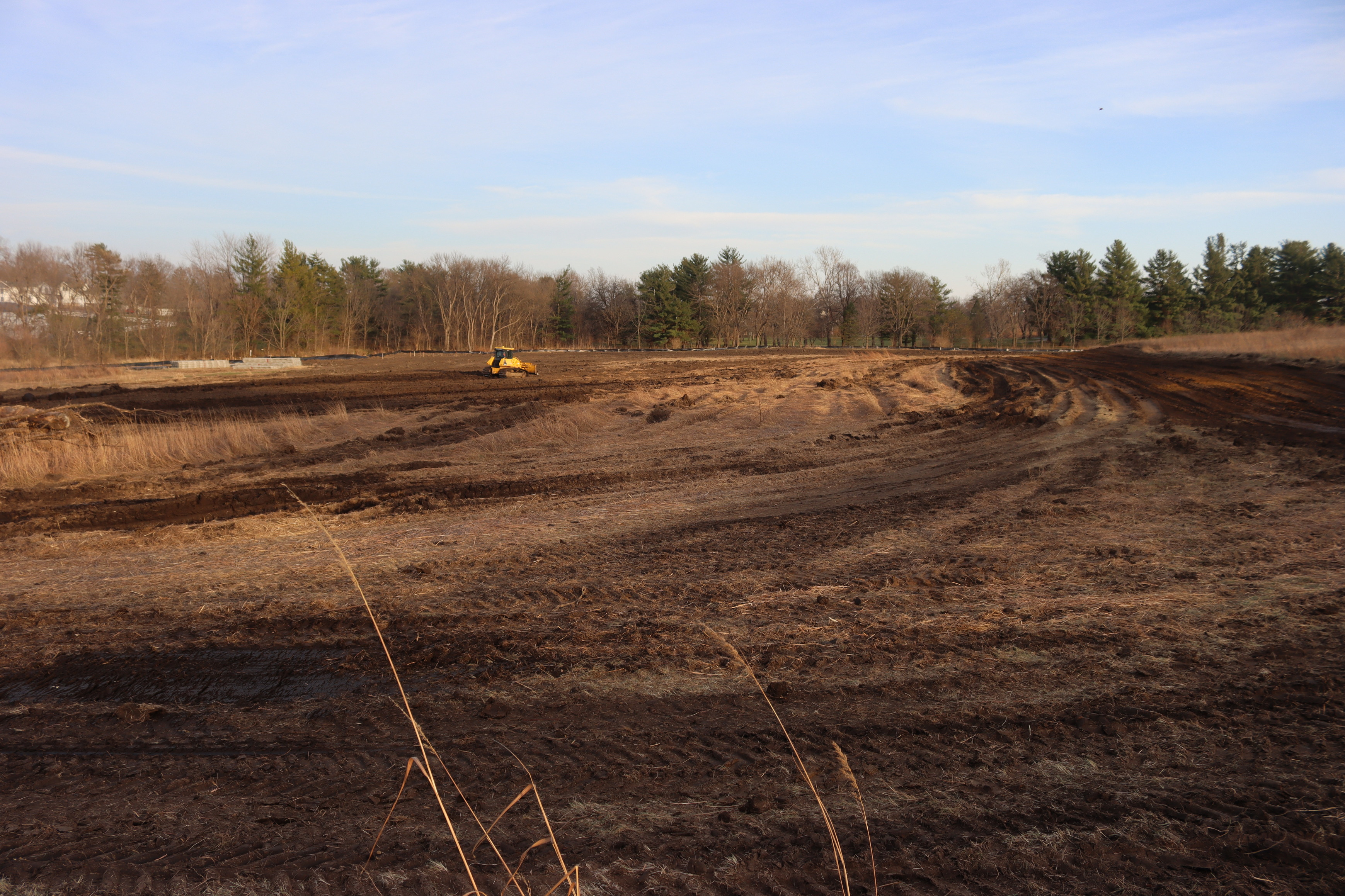 Excavated prairie area being turned into dention basin. A yellow bulldozer works just to the left of center. A temporary road runs along the right side of the image. Large tire tracks are visible in the foreground.