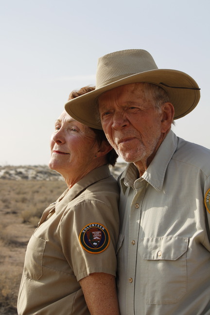 A man and a woman stand side to side dressed in the tan NPS volunteer uniform. The man is wearing a hat and looking towards the camera. The woman looks forward towards an eastern horizon. The desert unfolds in the background meeting white sand dunes covered in plants under a pale blue sky. 
