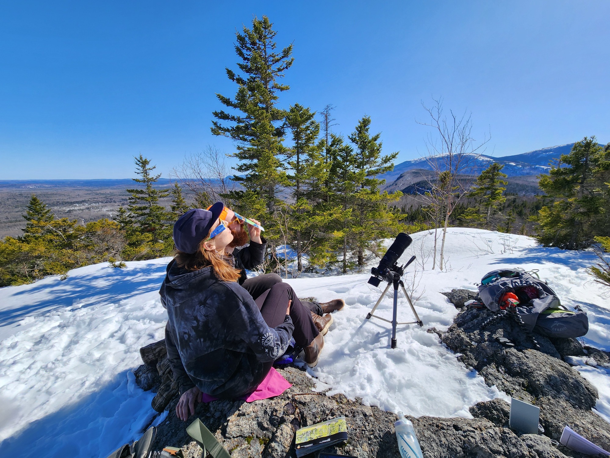 A woman and a man sit on top of a snow covered mountain to view the eclipse with solar shades and cameras. It is a sunny, clear, blue-sky day.