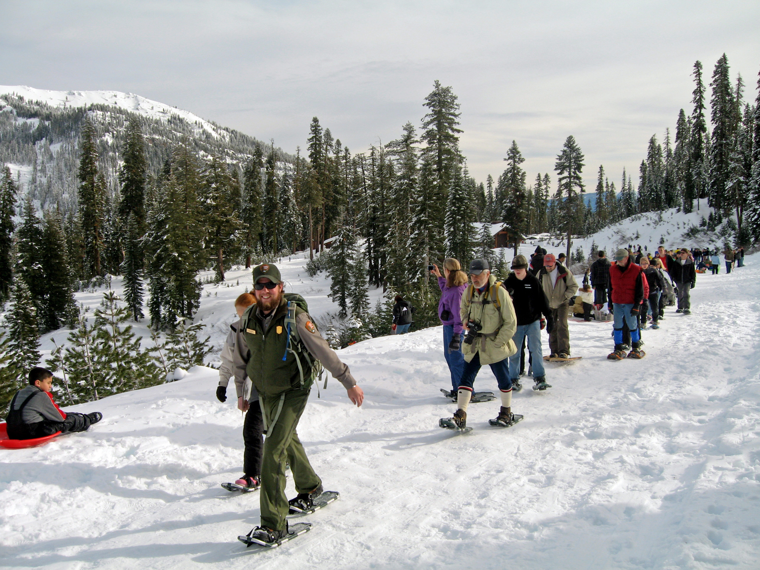a smiling park ranger leads a group of people of snowshoes on a snow-covered road.