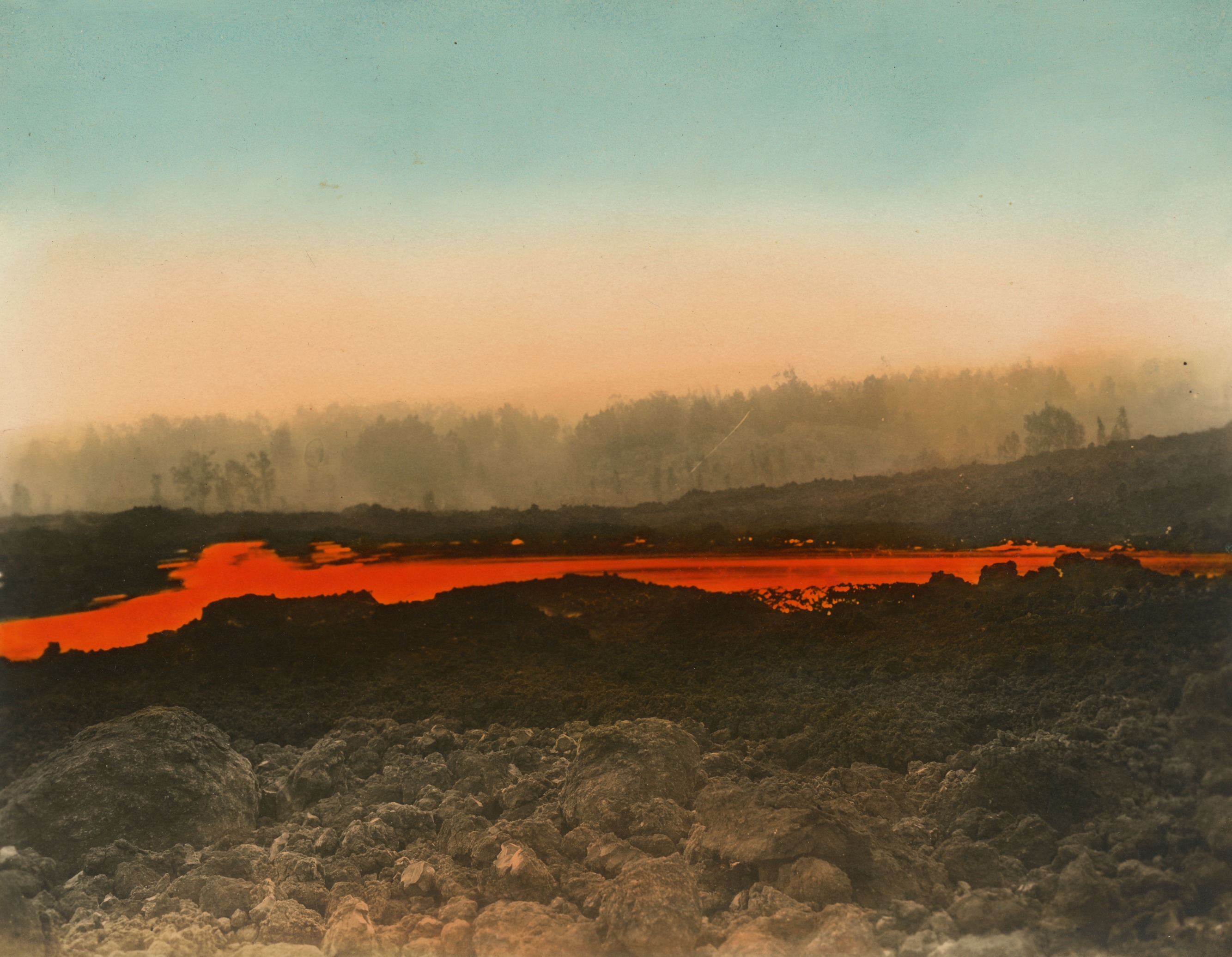 Hand-colored photograph of a landscape with a strip of molten lava. Originally a black and white photograph, the image has been colorized post-capture with bright red, orange, and light blue. The forefront of the image is comprised of large rocks and dried lava and has been tinted light orange. In the center of the image, molten lava flows horizontally, stretching from right to left, and has been colored bright red. The lava is travelling at an accelerated rate of speed, as evidenced by blurred lines as it splashes among the rocks. In the background, a dense tree line is seen, partially obstructed by the smoke from the eruption. The tree line and bottom part of the sky are tinted light orange, and the top half of the sky is light blue.