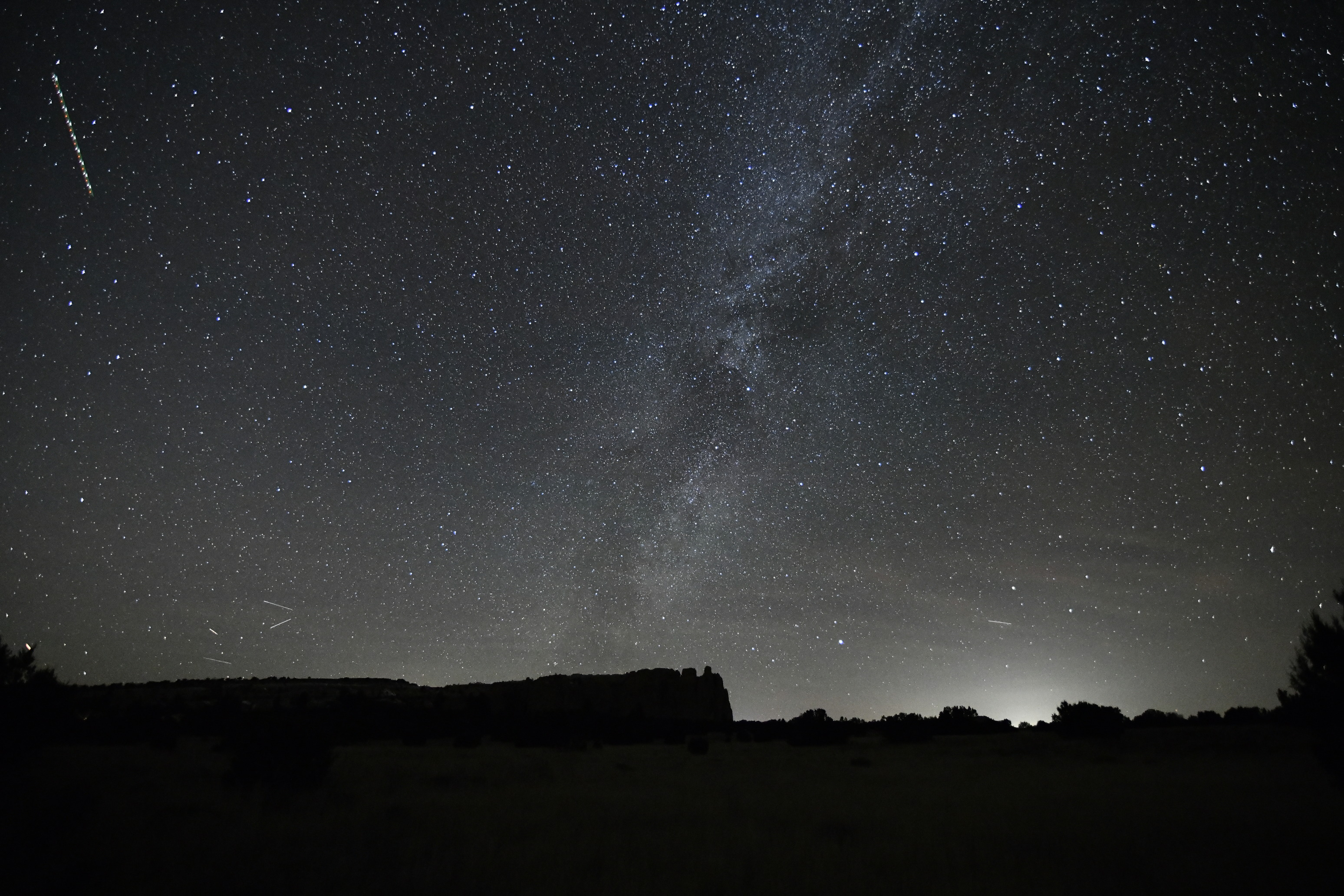 The Milky Way cuts across the sky behind the silhouette of a mesa. Light pollution from a nearby town glows in the lower right corner.