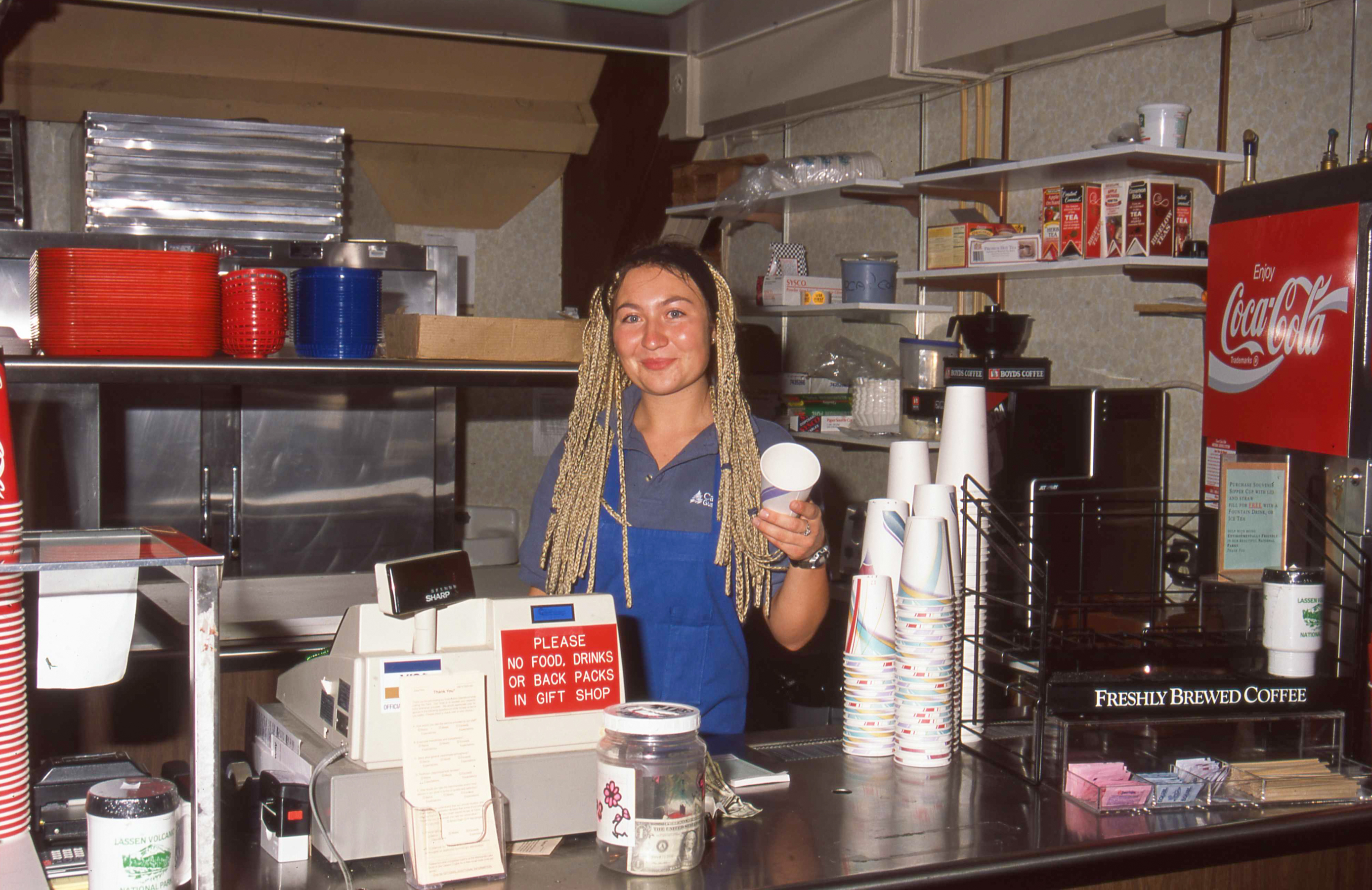 A young lady standing behind food service counter holding a cup.