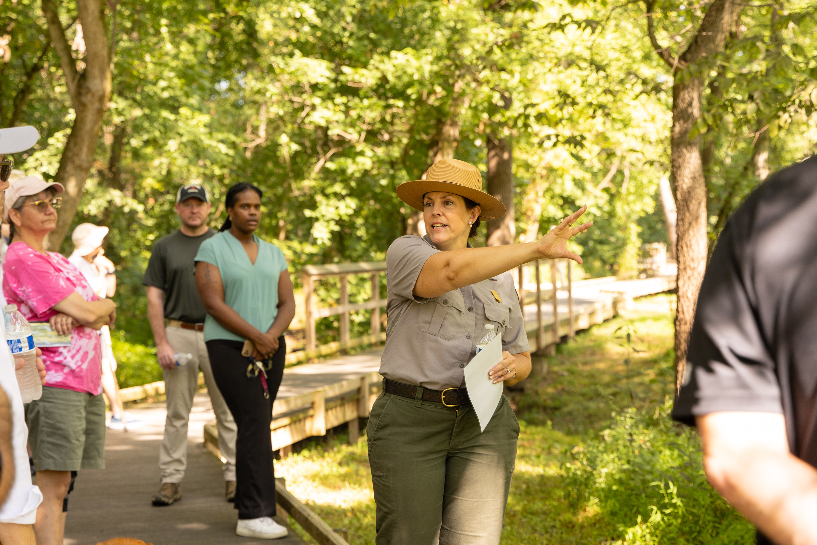 A park ranger in uniform gestures behind her to a patch of trees. Visitors surround her on a boardwalk. 